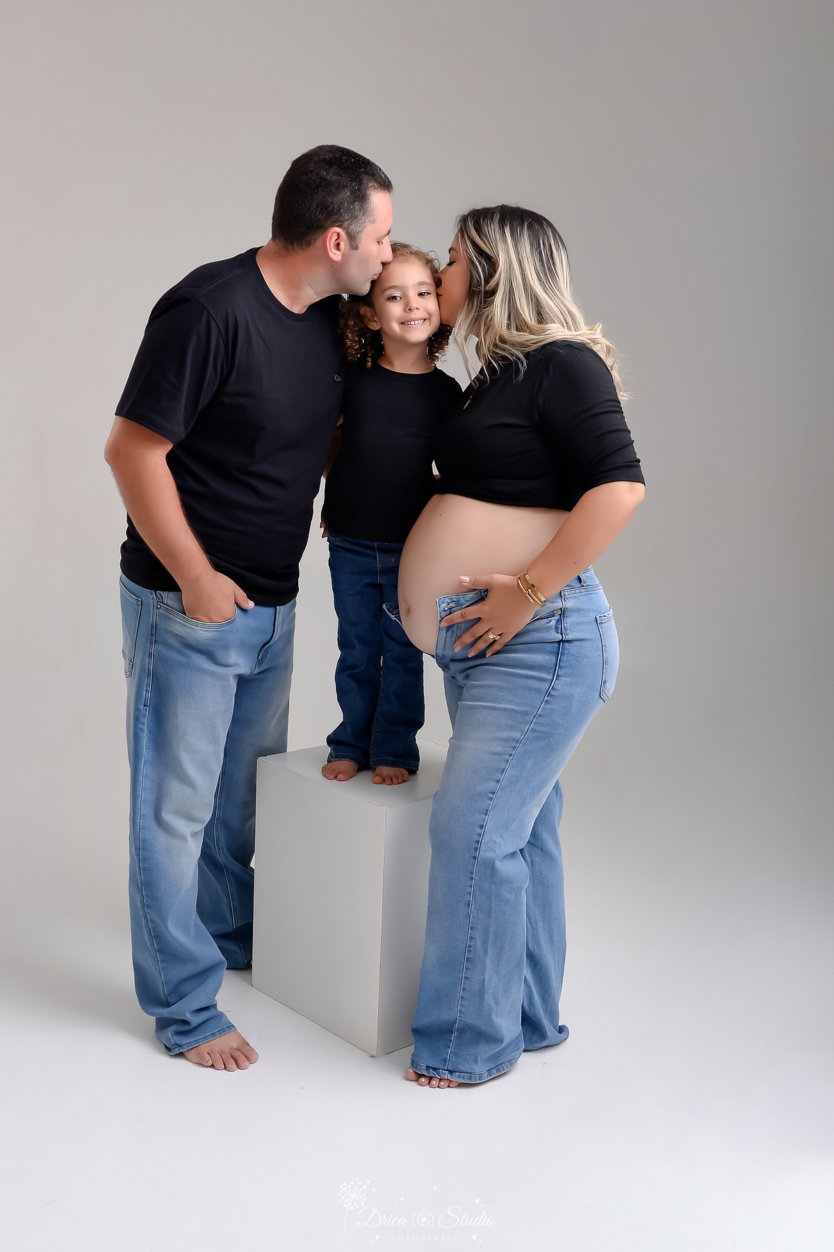 ensaio familia - ensaio gestante - mãe de 2 - irmã mais velha - ensaio em studio - xinguara -pará - familia usando çalça jeans e camisas pretas - filha mais velha sobre um banquinho - recebendo beijinho dos pais 