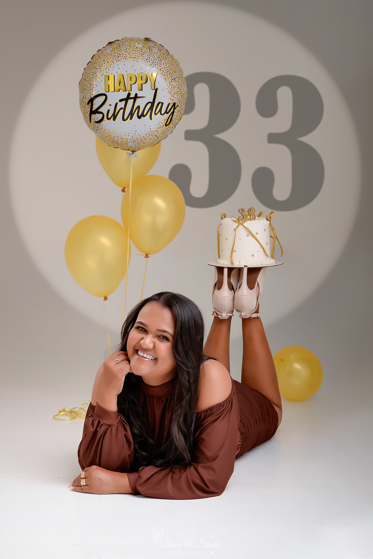 Aniversário - 33Anos - HappyBirthday - EnsaioFeminino -  EnsaioDeAniversário - aniversariante usando vestido marrom - deitada no chão - com bolo nos pés - com balões dourados - numero 33 ao fundo - sorrindo com uma das mãos no rosto -