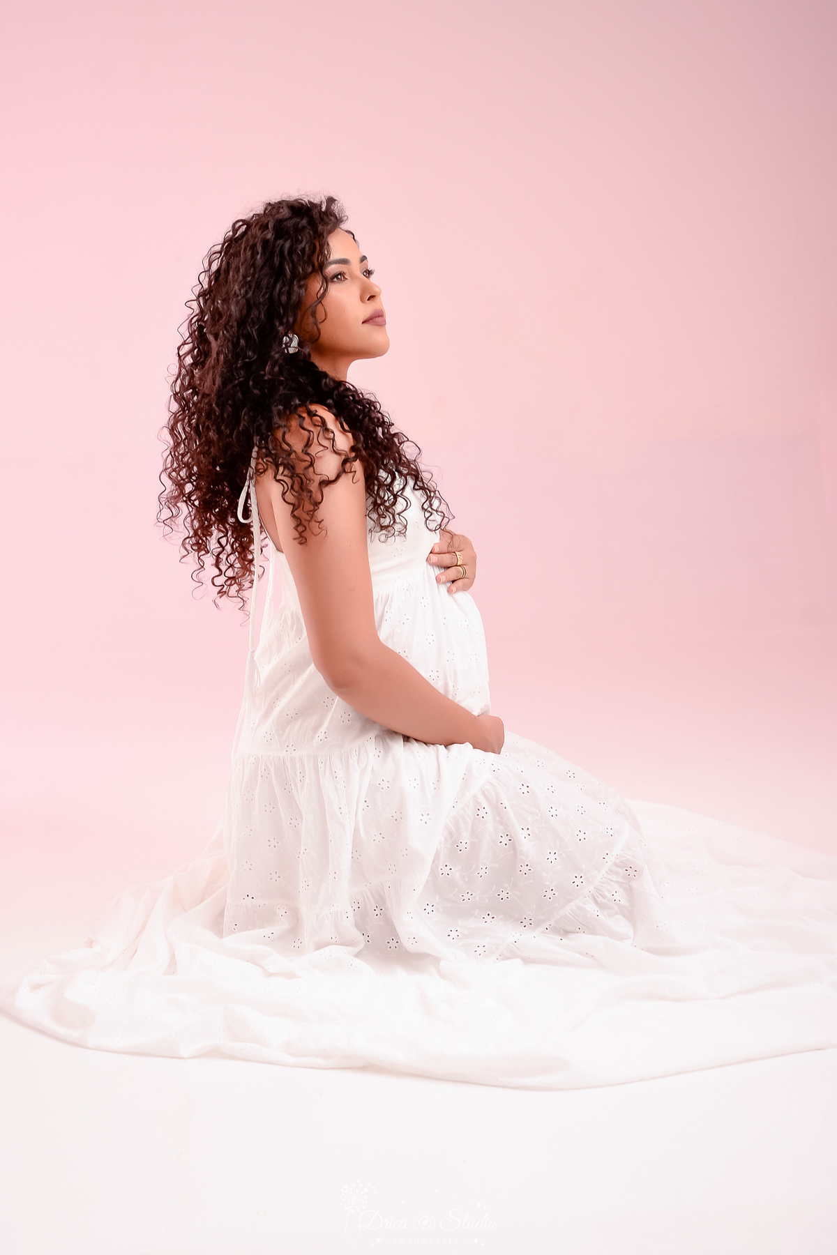 drica studio - ensaio gestante - mamãe de segunda viagem - usando vestido branco longo - sentada sobre o chão -  com as mãos na barriga - fundo com luz rosa - 