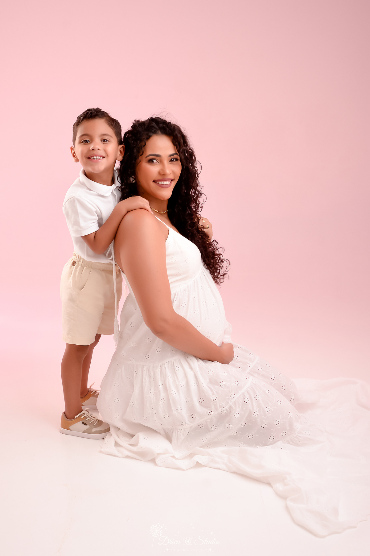 drica studio - ensaio gestante - mãe e filho - mamãe de segunda viagem - usando vestido branco longo - sendata sobre o chão - olhando para a camera e sorrindo - filho abraçando a mãe 