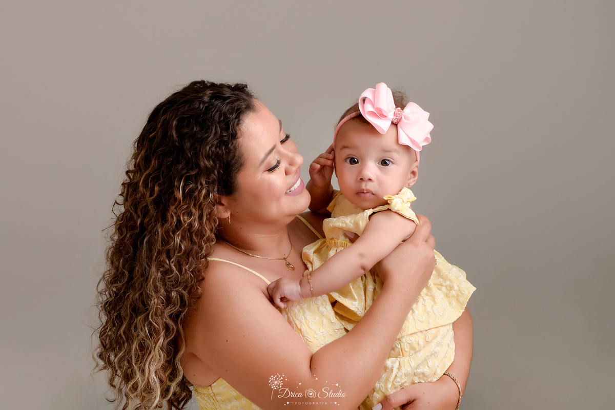Ensaio mãe e filha - amor incondicional - mãe e filha usando roupas iguais - tom amarelo - em pé com a filha nos braços - bebê usando laço rosa 