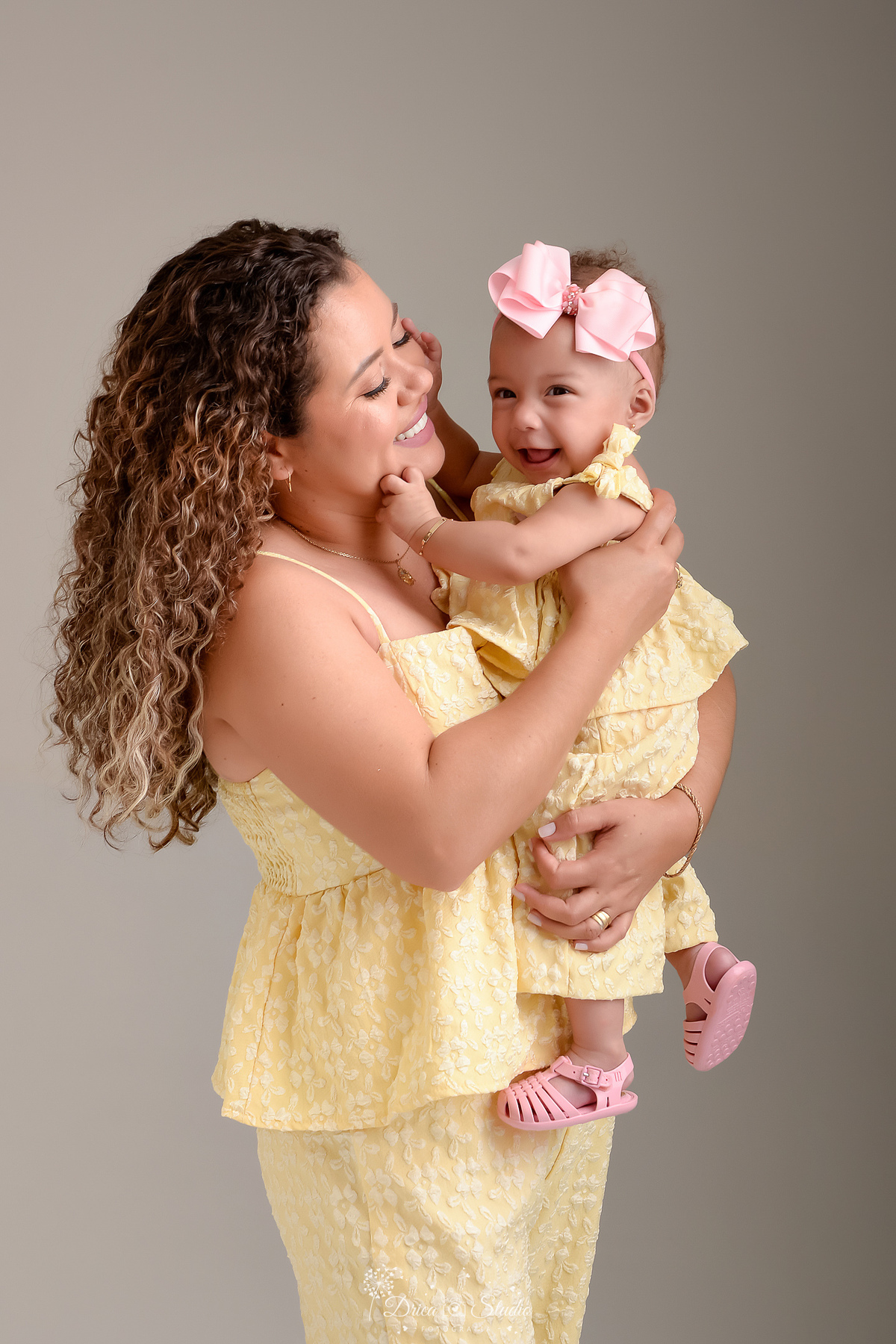 Ensaio mãe e filha - amor incondicional - mãe e filha usando roupas iguais - tom amarelo - em pé com a filha nos braços - bebê usando laço rosa -