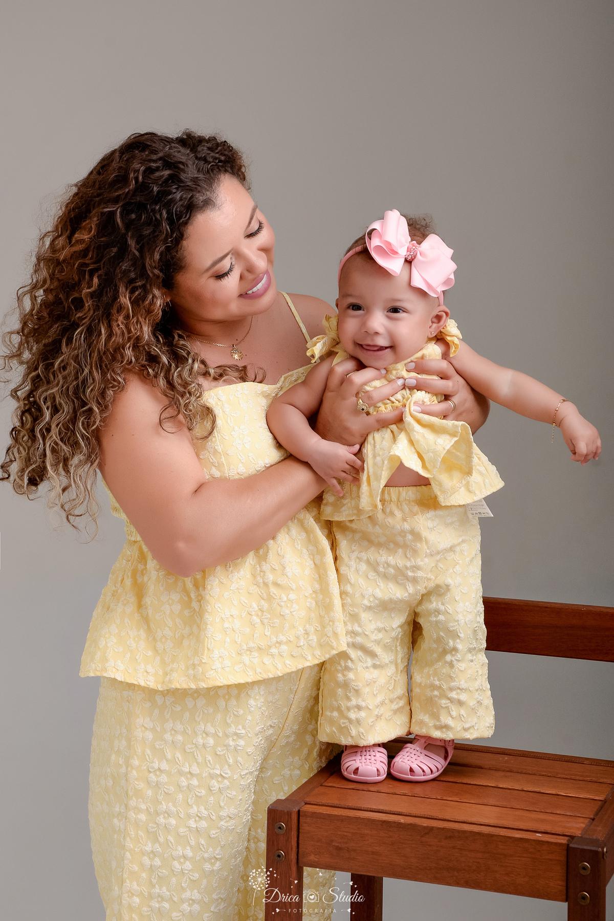 Ensaio mãe e filha - amor incondicional - mãe e filha usando roupas iguais - tom amarelo - em pé ao lado da filha que está sobre uma cadeira de madeira bebê usando laço rosa 