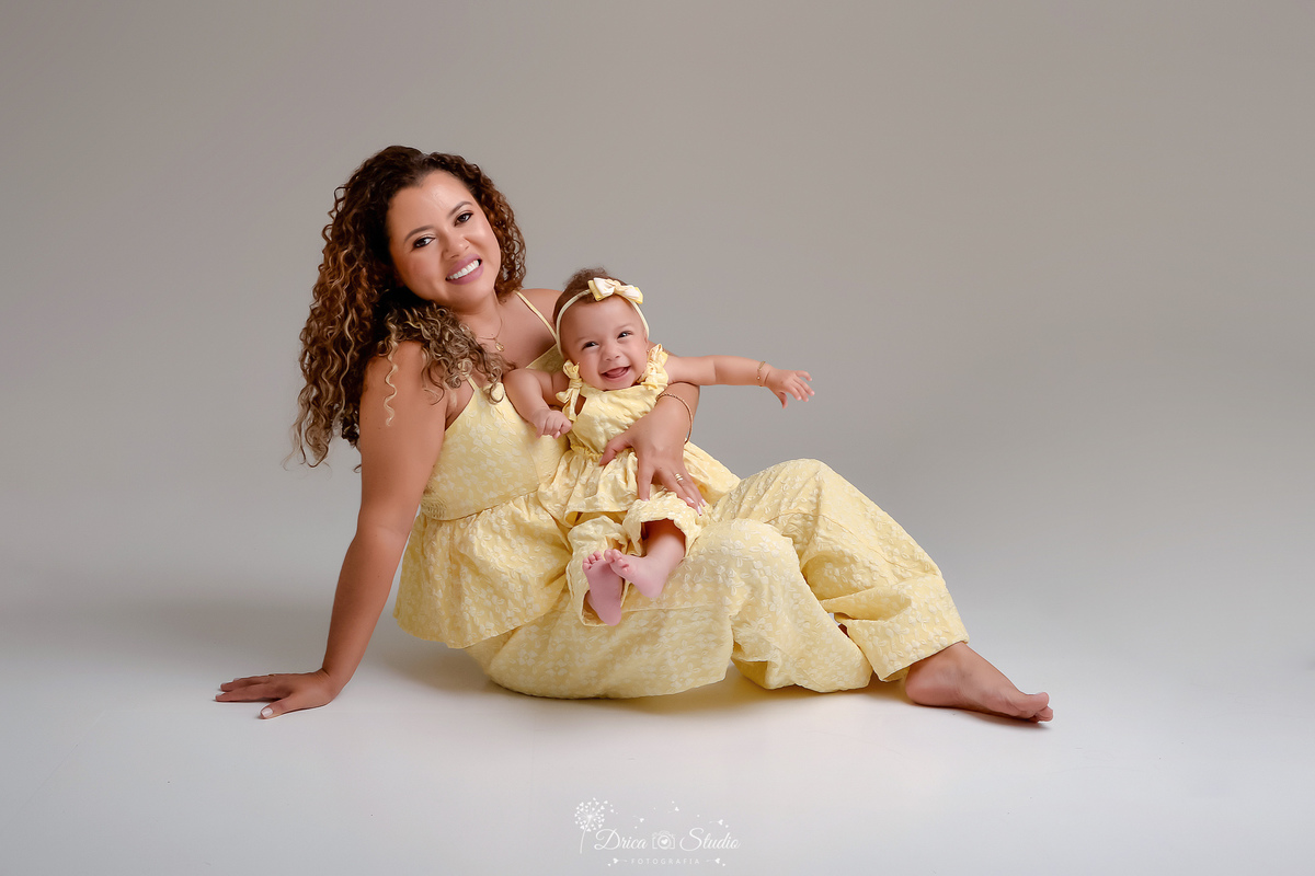 Ensaio mãe e filha - amor incondicional - mãe e filha usando roupas iguais - tom amarelo - sentada no chão com a filha no colo - bebê usando laço rosa - 