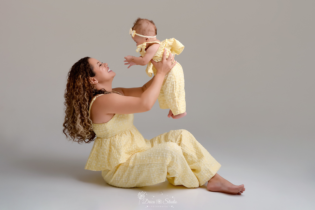Ensaio mãe e filha - amor incondicional - mãe e filha usando roupas iguais - tom amarelo- sentada no chão - brincando com a bebê - bebê usando laço rosa - 