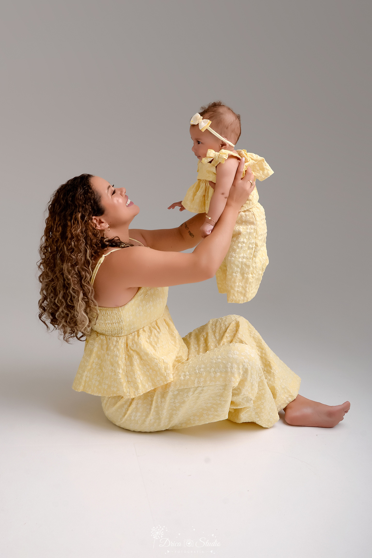 Ensaio mãe e filha - amor incondicional - mãe e filha usando roupas iguais - tom amarelo - sentada no chão - brincando com a bebê bebê usando laço rosa 