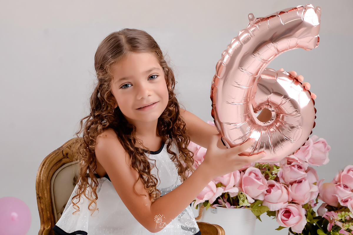 ensaio infantil - fotos de aniversario - 6 anos - menina - drica studio - xinguara - pará - foto em estudio - sentada em uma cadeirinha - segurando um balão com o numero da idade - ao fundo balões e flores rosas 