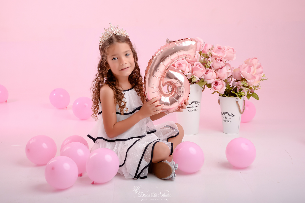 ensaio infantil - fotos de aniversario - 6 anos - menina - drica studio - xinguara - pará - sentada no chão - segurando um balão com o numero da idade - ao lado de dois vasos de flores rosas - balões rosas pelo chão - foto em estudio - fundo rosa 