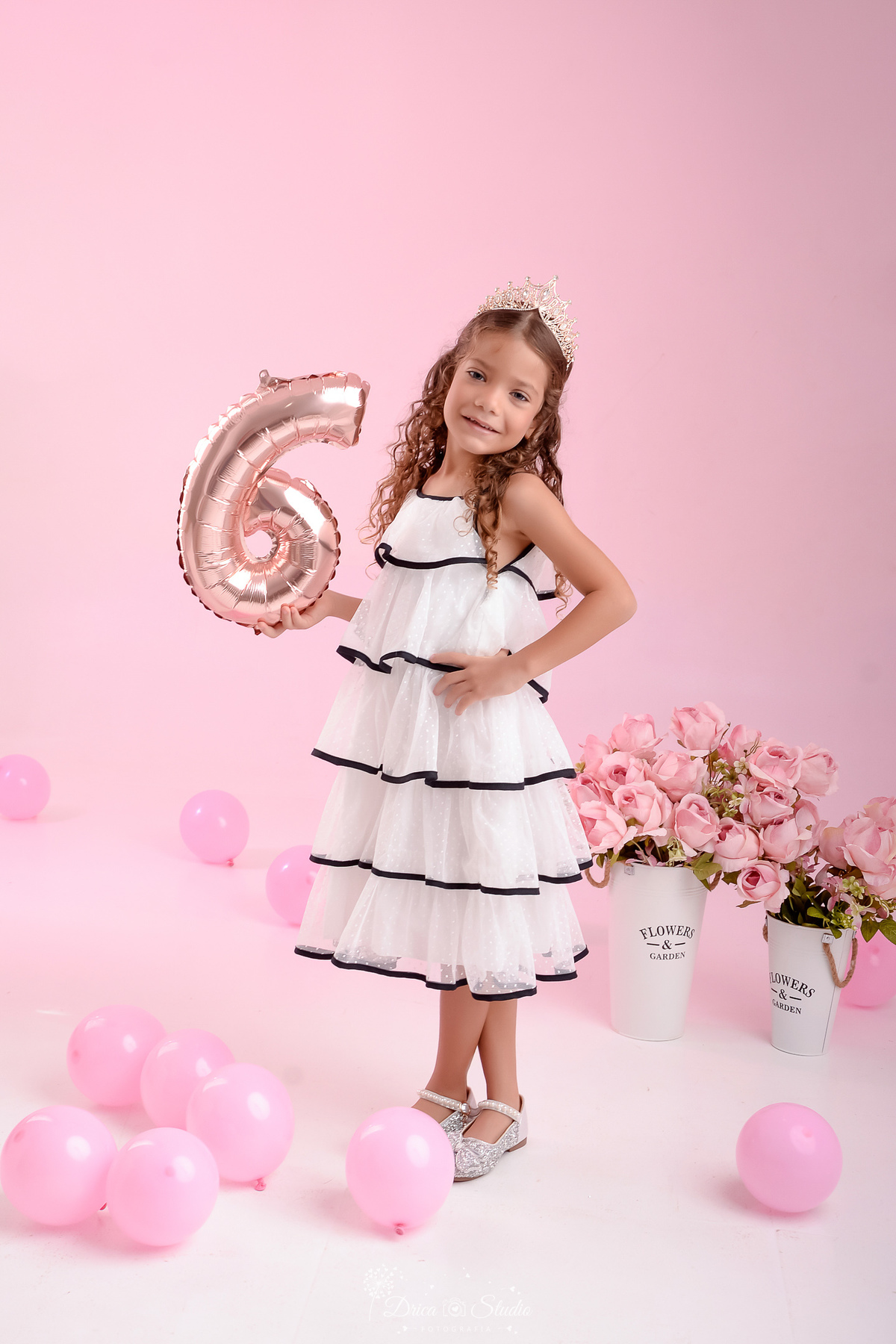 ensaio infantil - fotos de aniversario - 6 anos - menina - drica studio - xinguara - pará - segurando um balão com o numero da idade - usando vestido branco com detalhe preto - fundo rosa - flores rosas - balões rosas - foto em estudio -