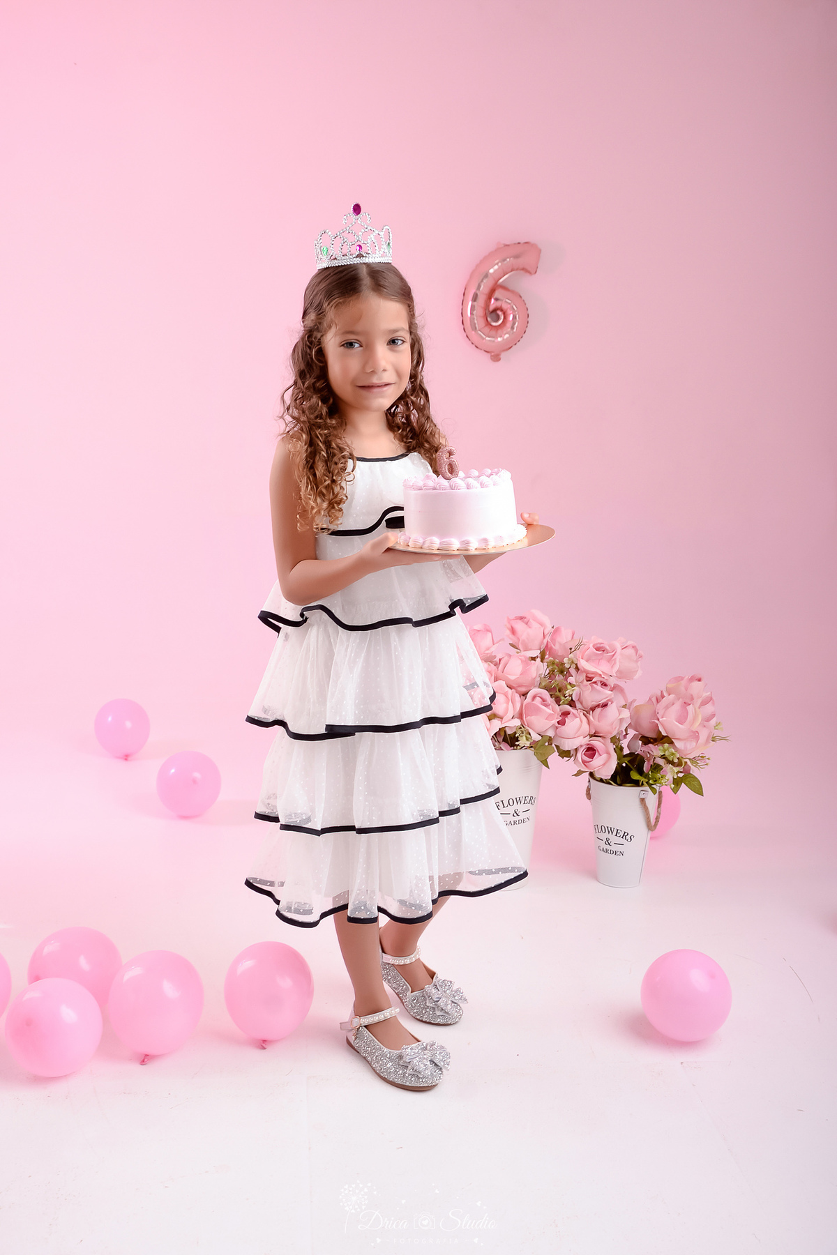 ensaio infantil - fotos de aniversario - 6 anos - menina - drica studio - xinguara - pará - foto em estudio - fundo rosa - balões rosas - flores rosas - usando vestido branco com detalhe preto - segurando um bolo - balão de numero 6 ao fundo 