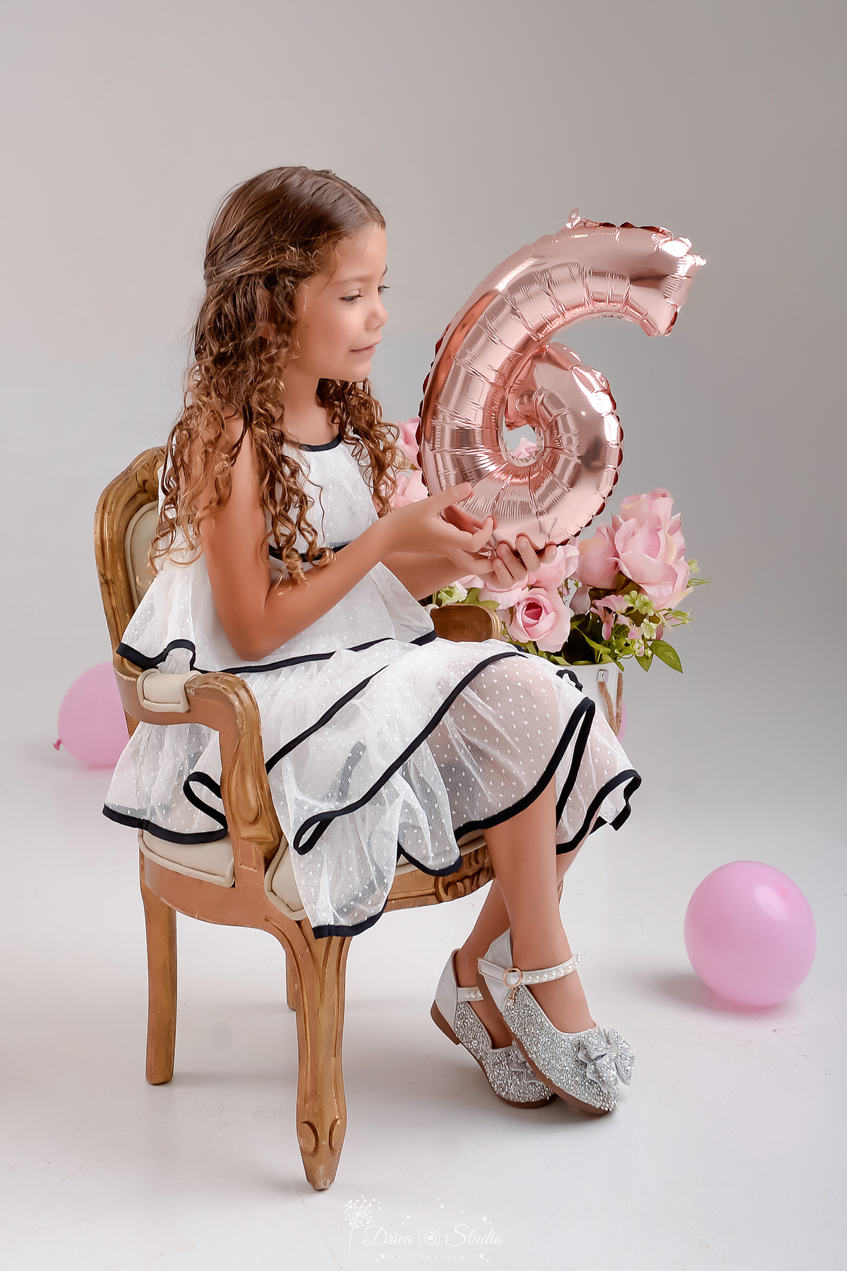 ensaio infantil - fotos de aniversario - 6 anos - menina - drica studio - xinguara - pará - sentada em uma cadeirinha - segurando o balão com o numero da idade - balões e flores rosas ao fundo - foto em estúdio 