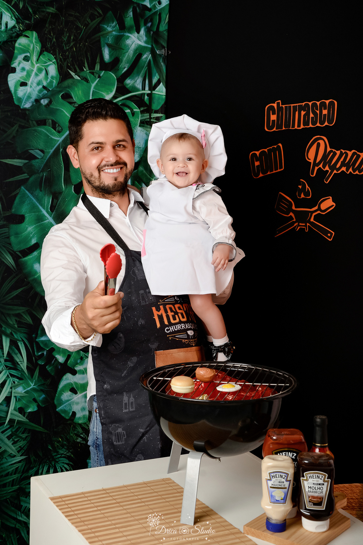 especial dia dos pais - cenario de churrasco - com churrasqueira preta - molhos de churasco - pai e filha usando aventais de churrasco - fundo com folhagens verde - lchurras com o papai escrito na parede na cor preta e laranja 