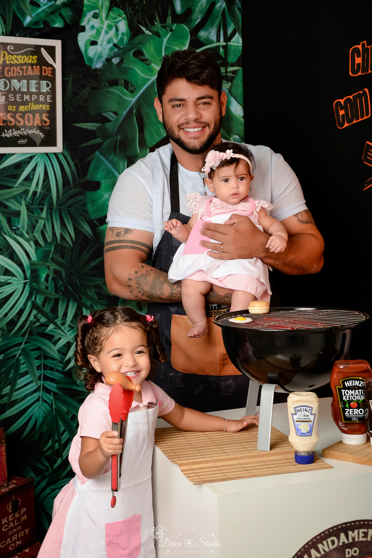 especial dia dos pais - pai e filhas - necario de churrasco - com churrasqueira preta - filhas usando vestidos rosas e avental - pai usando avental de churrasco - molhos de churrasco sobre a bancada - fundo com folhagems verdes 