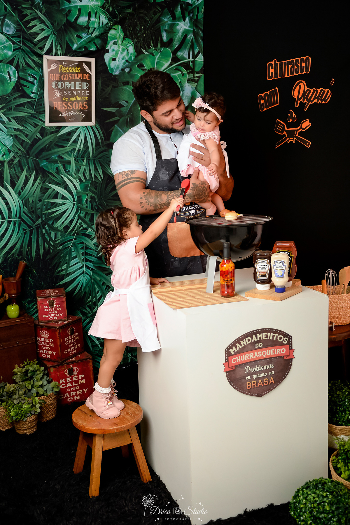 Especial dia dos pais - cenario de churrasco com o papai - pai e filhas - cenario com churrasqueira preta - filhas usando vetidos rosas e aventais de churrasco - fundo com folhagems verdes de um lafo e do outro lado preto 