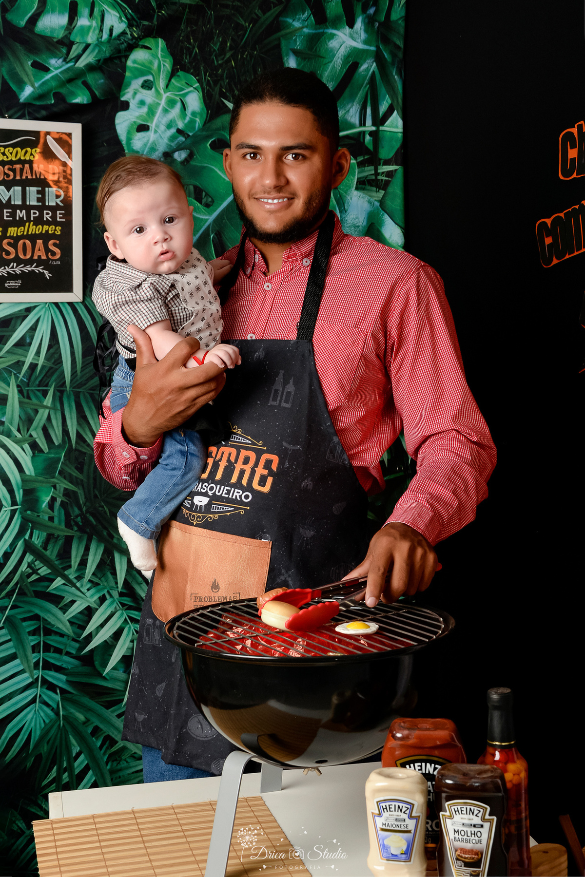 especial dia dos pais - cenario de churrasco - churrasqueira preta - pai usando avental de churrasco preto - fundo com folhagem verde - acompanhamento mensal - drica studio - xinguara - pará - pai e filho 