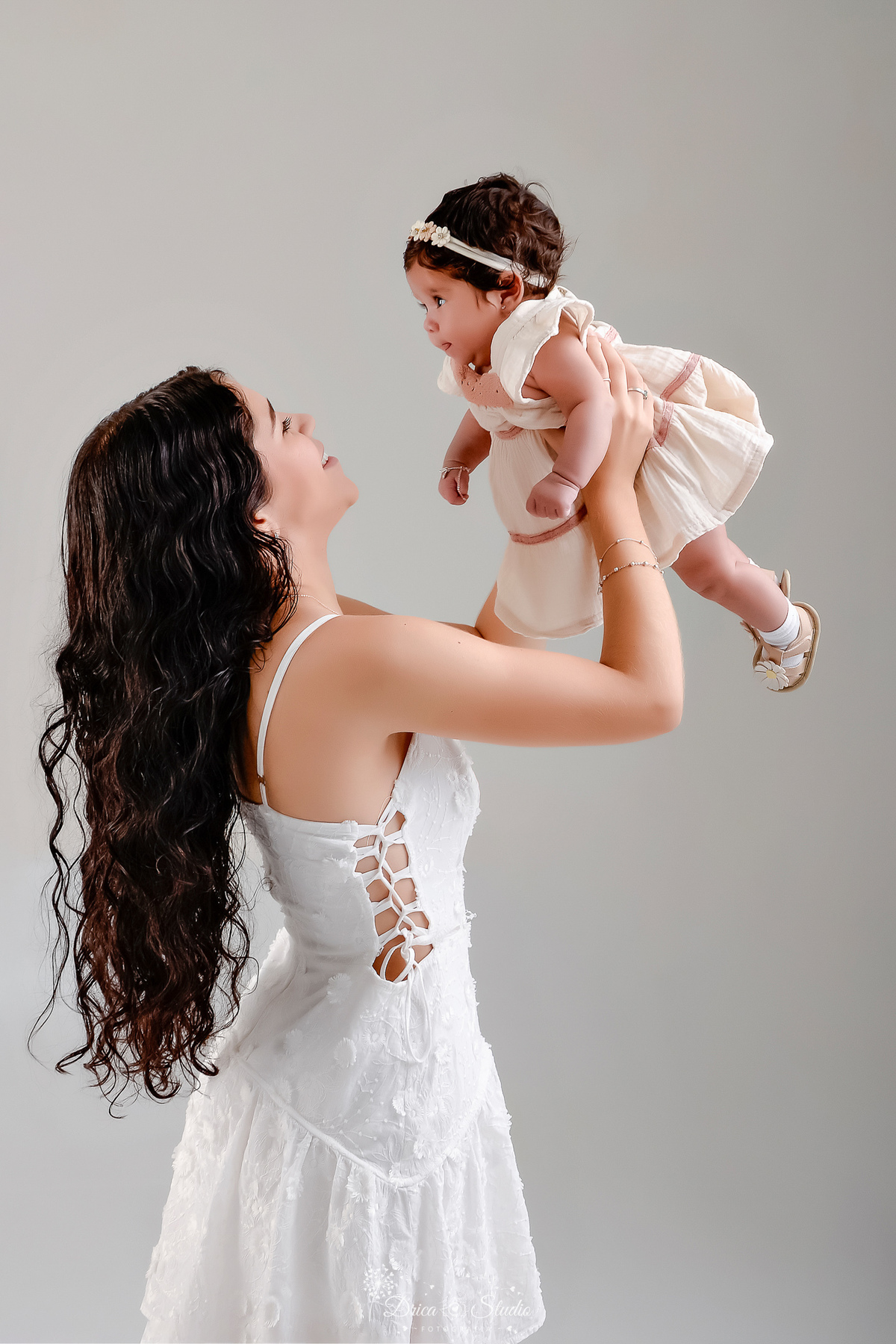 mãe e filha - acompanhamento mensal - fotos em estúdio - drica studio - xinguara - pará - mãe usando vestido branco - erguendo a filha - filha usando bestido bege - fundo branco - mãe e filha se olhando -