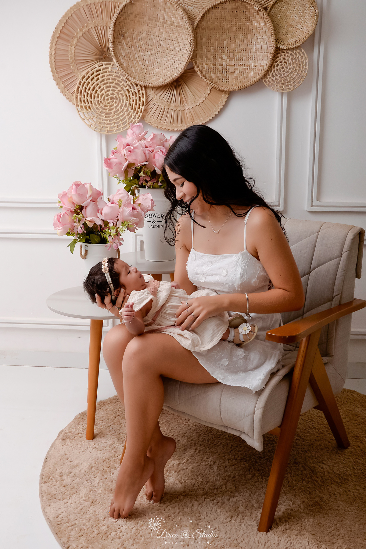 mãe e filha - acompanhamento mensal - fotos em estúdio - drica studio - xinguara - pará - mãe sentada em uma poltrona - com a filha no colo - ao lado duas mesinhas com arranjo de flores rosas - mãe usando vestido branco - olhando e sorrindo para a filha 