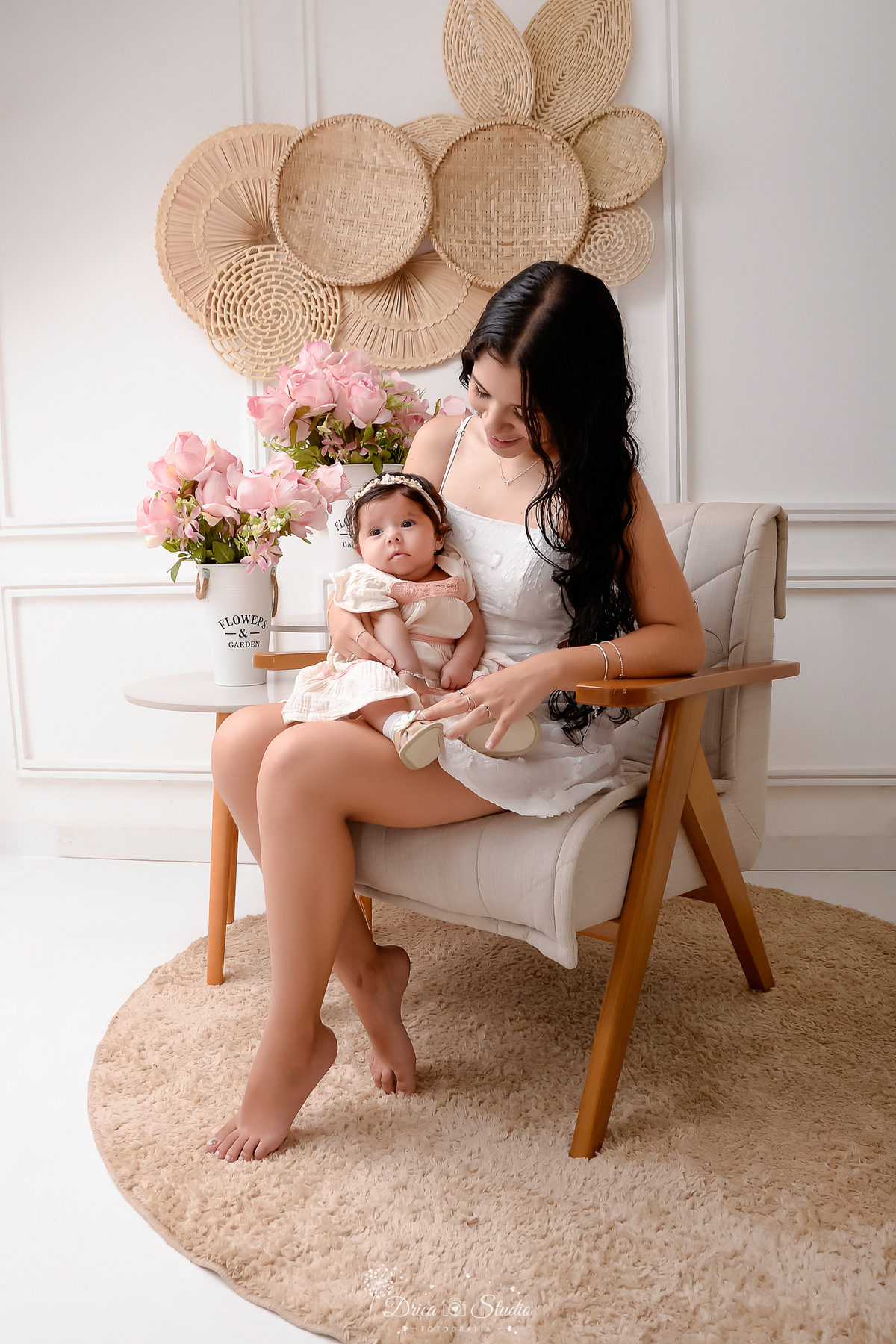 mãe e filha - acompanhamento mensal - fotos em estúdio - drica studio - xinguara - pará - mãe sentada em uma poltrona - ao lado duas mesinhas com arranjo de flores rosas - mãe usando vestido branco - segurando a filha em seu colo 