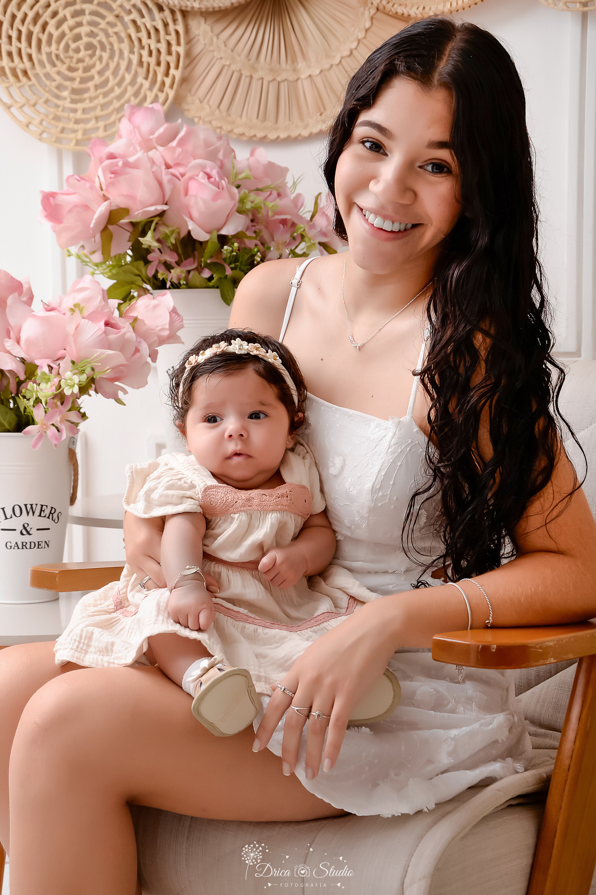 mãe e filha - acompanhamento mensal - fotos em estúdio - drica studio - xinguara - pará - mãe sentada em uma poltrona - ao lado dois arranjos de flores rosas - mãe usando vestido branco - segurando a filha no colo - 