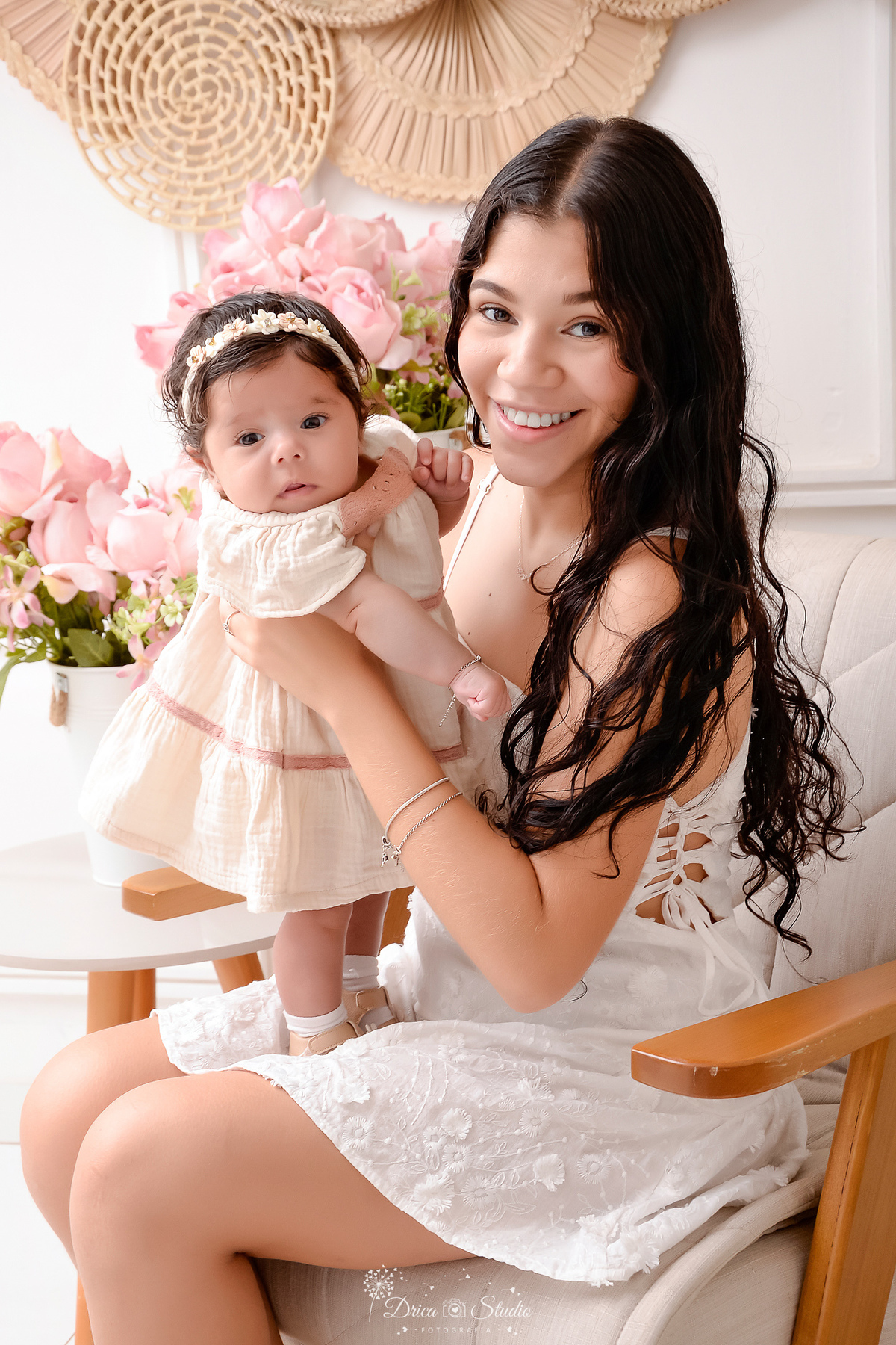 mãe e filha - acompanhamento mensal - fotos em estúdio - drica studio - xinguara - pará - mãe sentada em uma poltrona - segurando sua filha no colo - ao lado dois arranjos de flores rosas - filha usando vestido bege e laço 