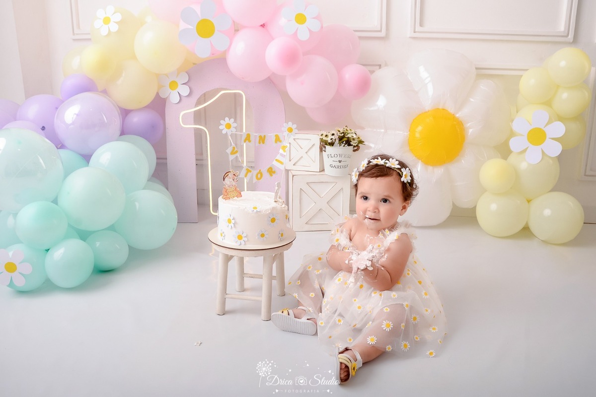 drica studio - smash the cake - 1 aninho - fim de acompanhamento mensal - bebe sentada no chão ao lado do bolo - cenário de margaridas com balões em tons pastéis - 1 de led - bolo branco com margaridas - bebe usando vestido branco 