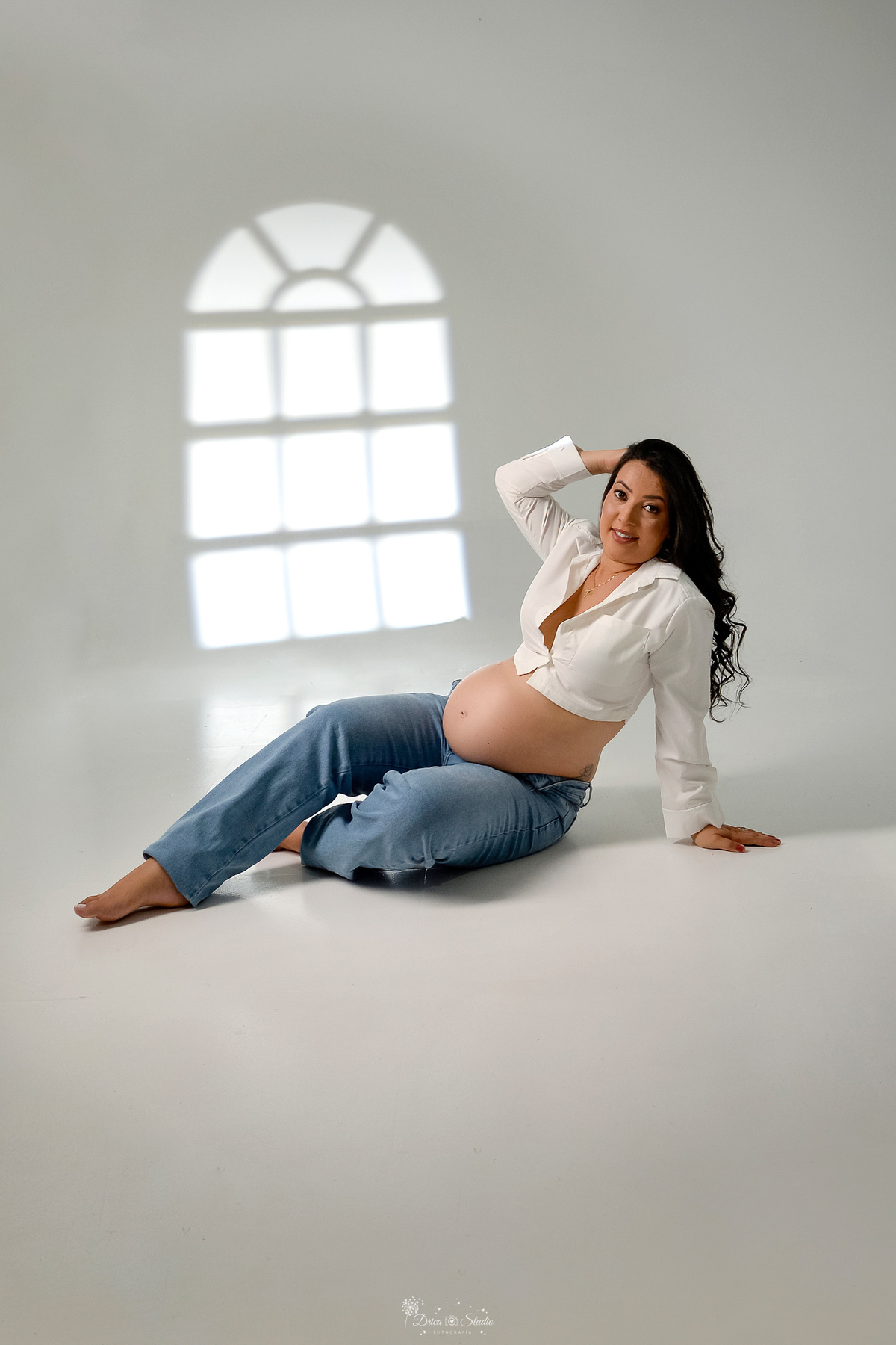 -ensaiogestante -fotografiagestante  -gravidez -maternidade - gestante sentada o chão - usando calça jeans e camisa branca com uma das mãos no cabelo - luz de fundo branca 