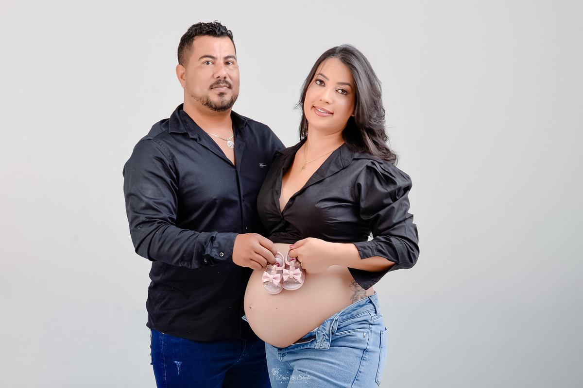 -ensaiogestante -fotografiagestante  -gravidez -maternidade - casal usando calça jeans e camisa preta - com sapatinho da bebê sobre a barriga - fundo branco 