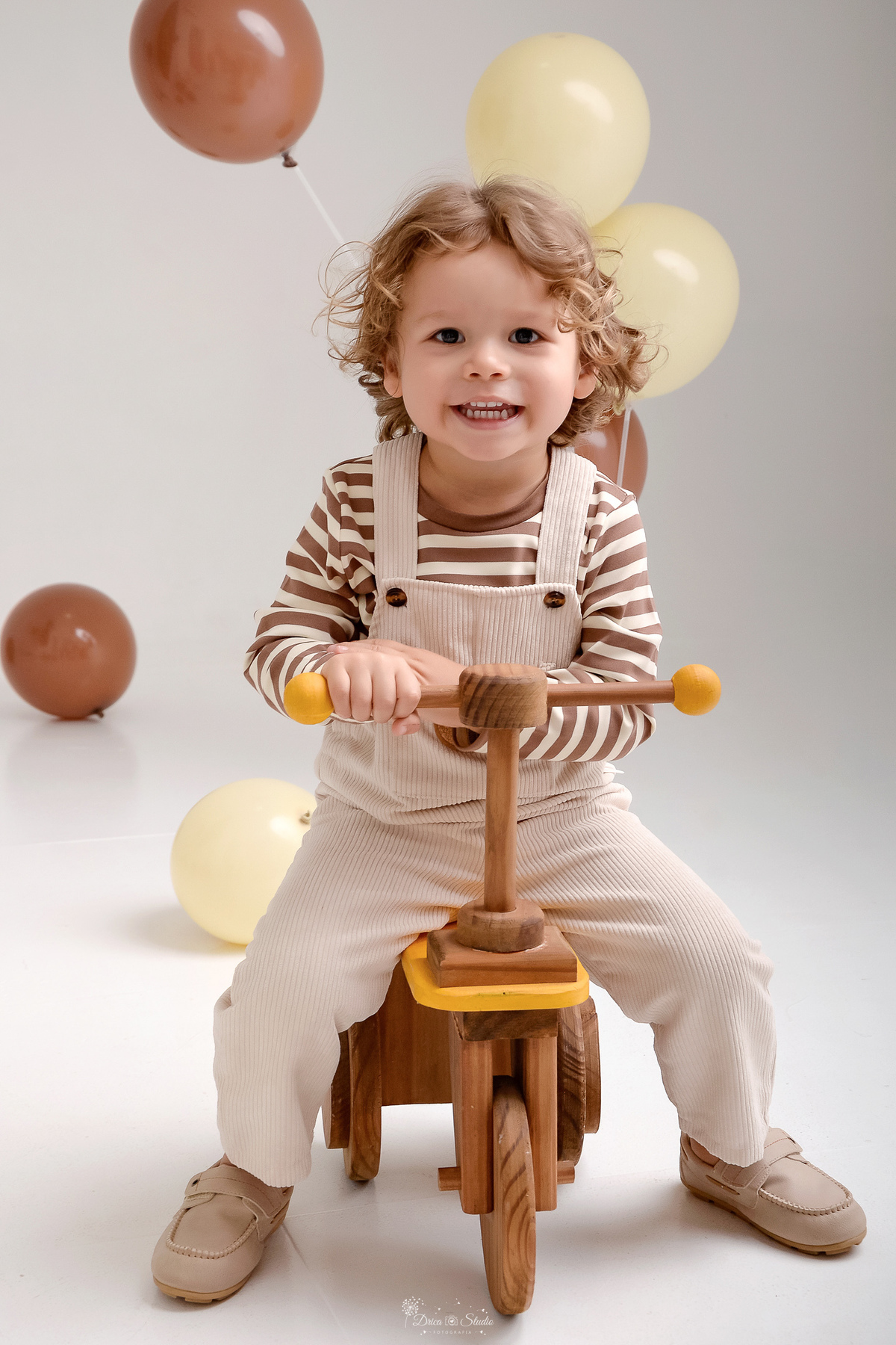 ensaio de aniversario - bebê de 2 anos - ensaio em estudio - drica studio - xinguara - pará - bebê em uma motinha de madeira - usando macacão bege com camisa listrada - ao fundo balões no chão na cor marrom e bege 