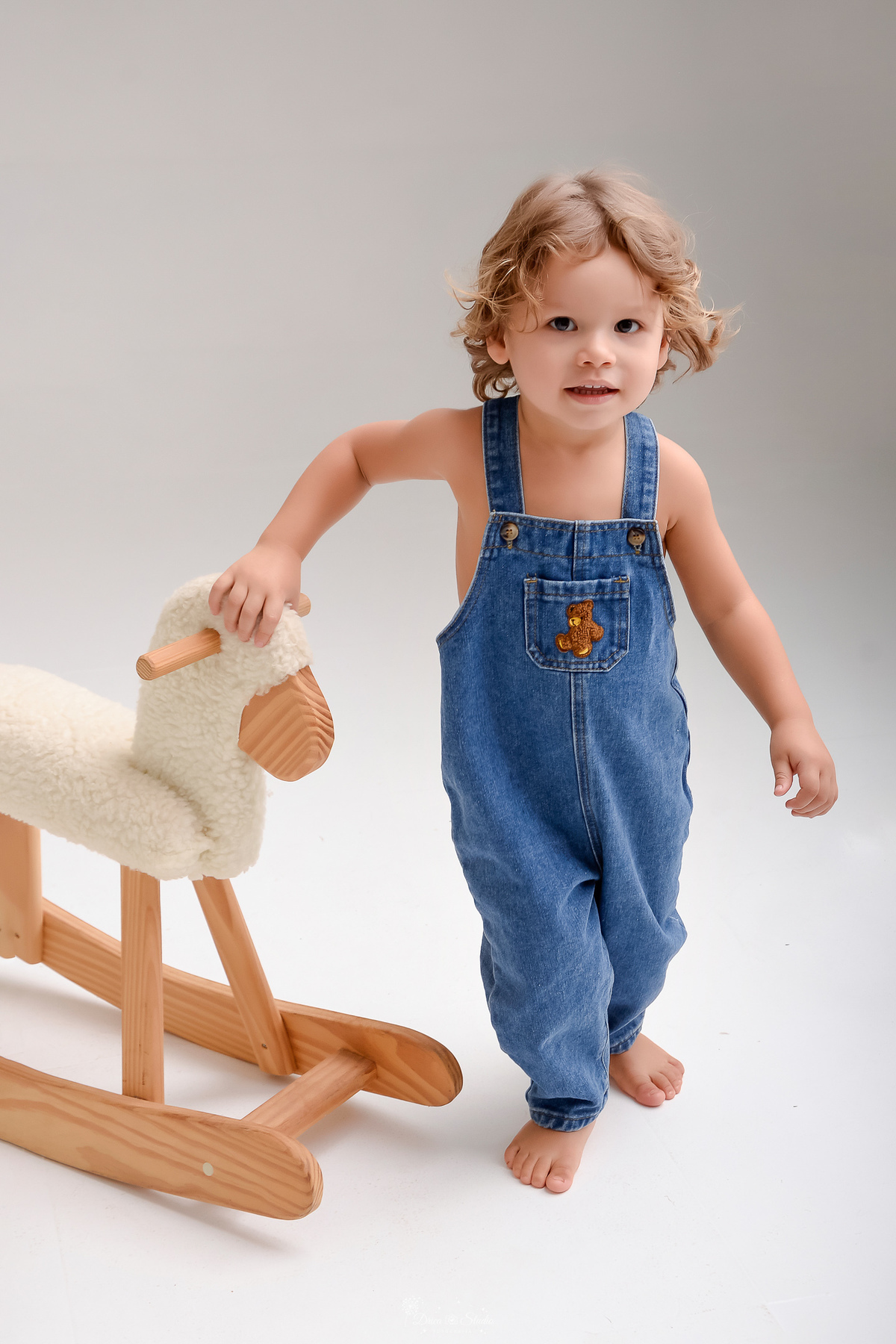 ensaio de aniversario - bebê de 2 anos - ensaio em estudio - drica studio - xinguara - pará - bebê usando macacão jeans - com o desenho de um ursinho marrom - com um cavalinho de madeira branco - 
