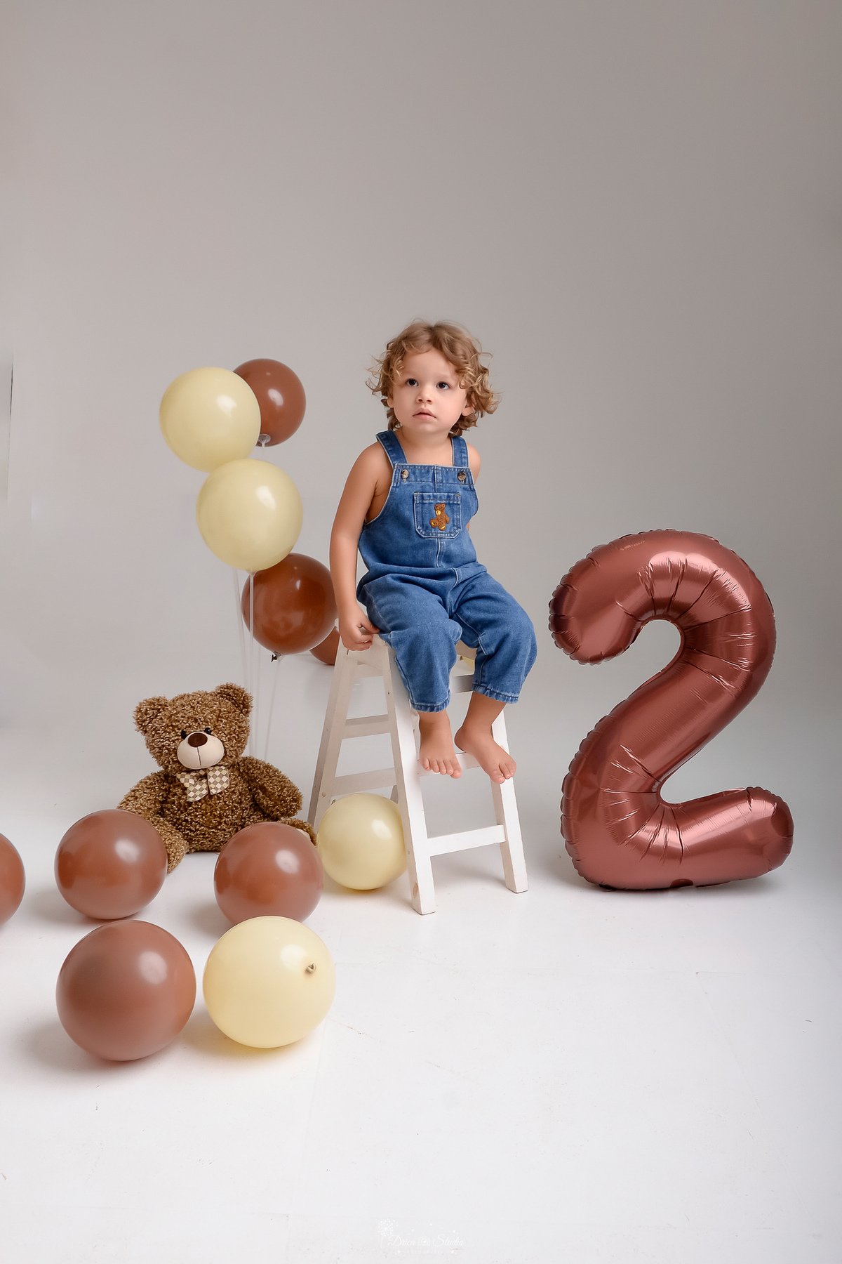 ensaio de aniversario - bebê de 2 anos - ensaio em estudio - drica studio - xinguara - pará - bebe sentado em uma escadinha branca -  valão de numero 2 marrom - balãos marrons no chão - ursinho de pelucia marrom no chão - usando macacão jeans 
