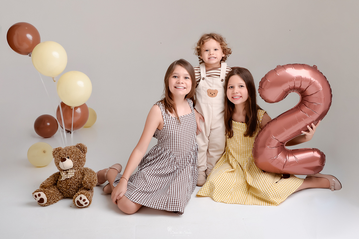 ensaio de aniversario - bebê de 2 anos - ensaio em estudio - drica studio - xinguara - pará - balão de numero 2 marrom - irmãs sentadas no chão - irmã mais velha segurando o balão de numero 2 ursinho marrom no chão - irmão abraçando as irmãs 