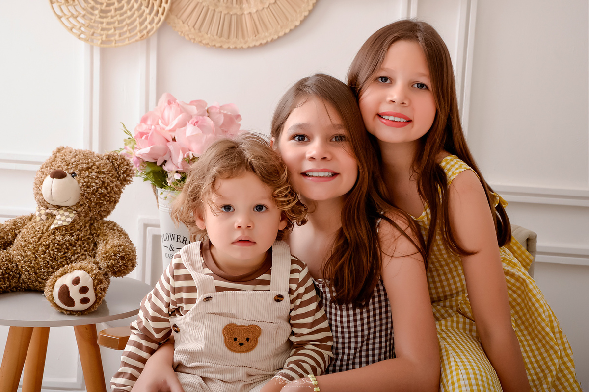 ensaio de aniversario - ensaio em familia - irmãs mais velhas - irmã mais velha sentada em uma poltrona - coom irmão mais novo no colo - irmã do meio ao lado - ursinho de pelucia marrom ao lado - 