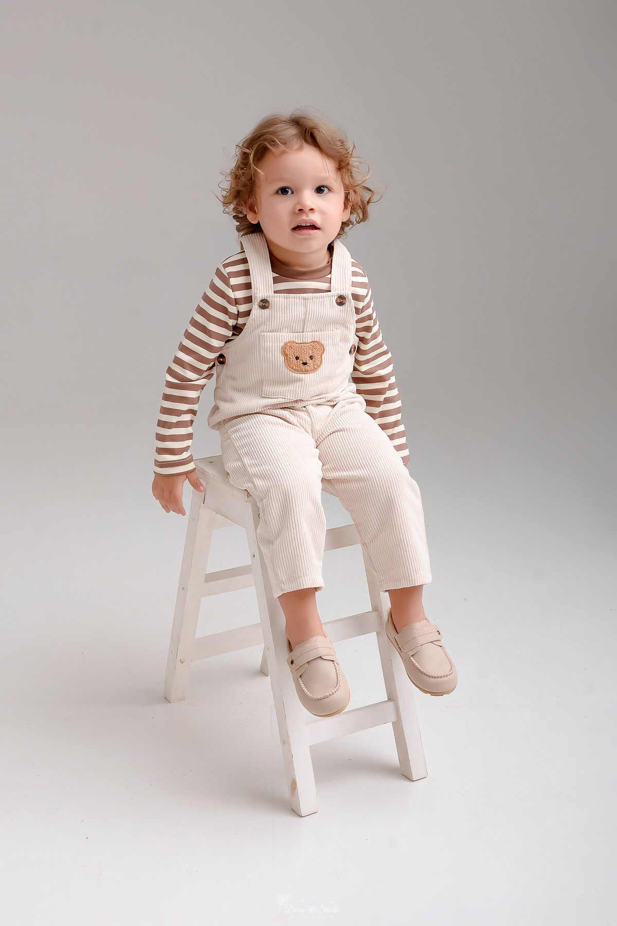 ensaio de aniversario - bebê de 2 anos - fotos em estudio - fotos em fundo branco - bebê sentado em um baquinho branco - usando macacão claro com camisa listrada - no tema de ursinho 