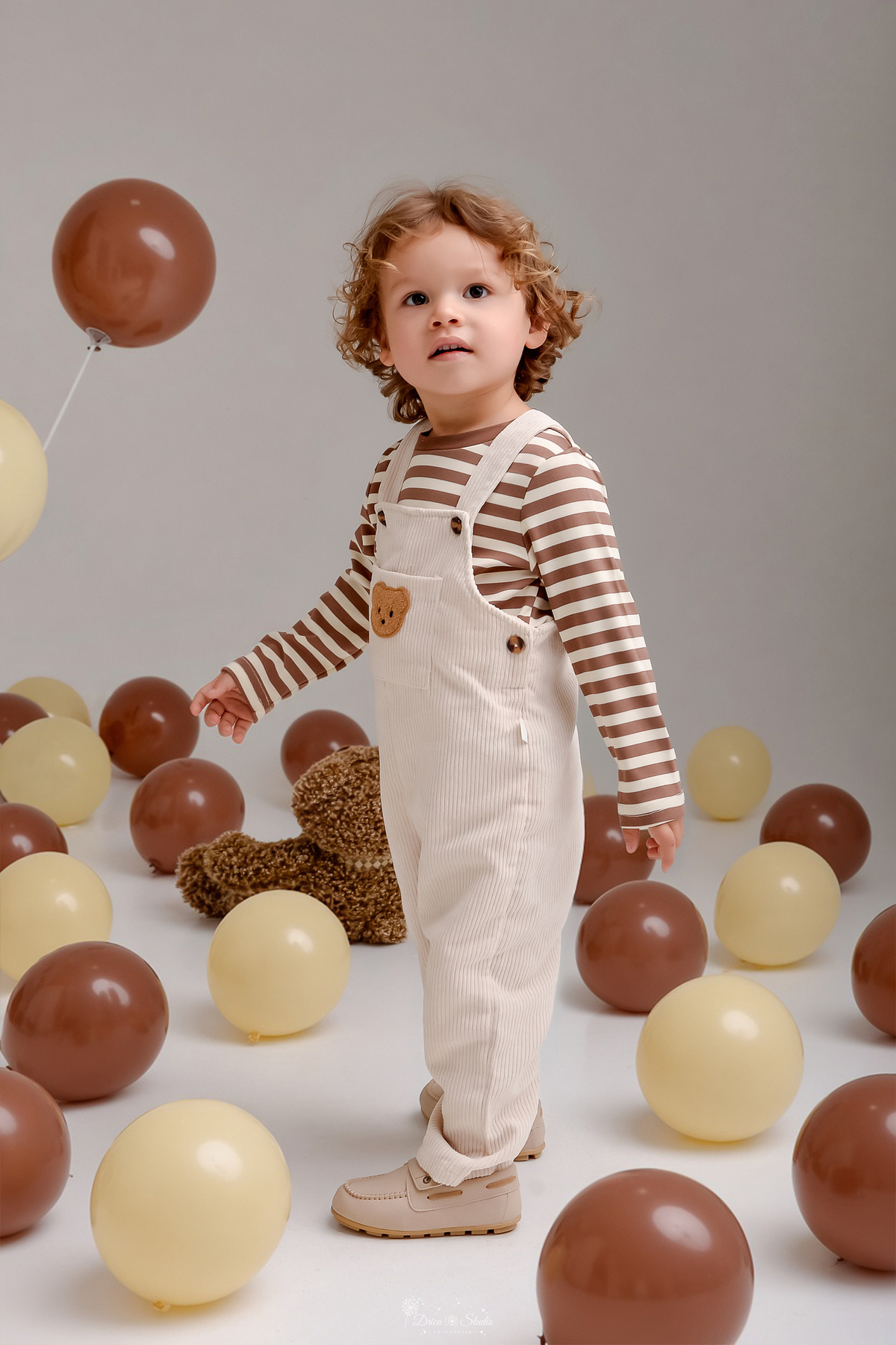 aniversário de 2 anos - fotos de aniversario - ensaio em estudio - bebê usando macacão claro - com camisa listrada - tema ursinho - ao chão balões nas cores marrom e bege - ursinho de pelucia marrom ao chão 