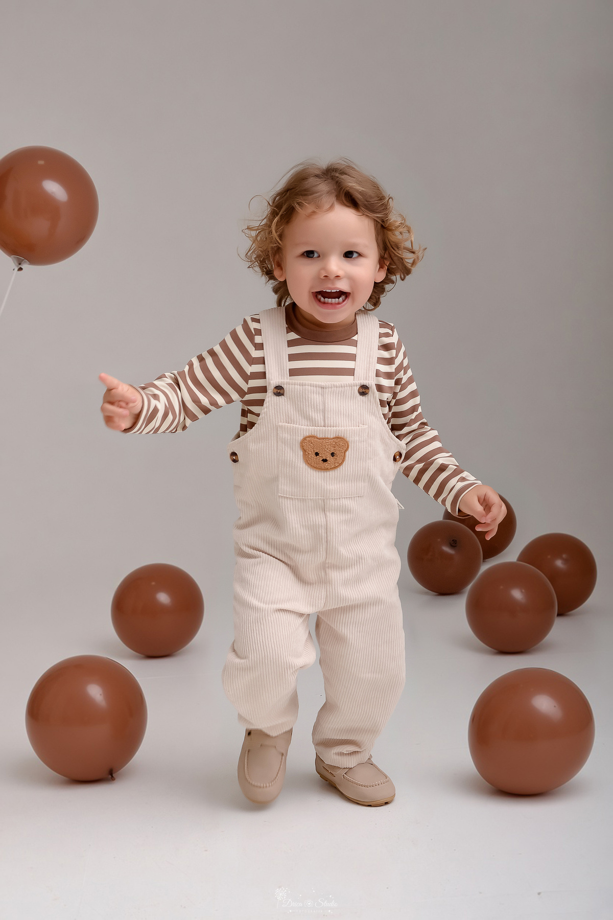 ensaio de aniversario - bebê de 2 anos - ensaio em estudio - drica studio - xinguara - pará - bebê em pé - balões marrons pelo chão -  usando macacão claro com camisa listrada - tema ursinho marrom - 