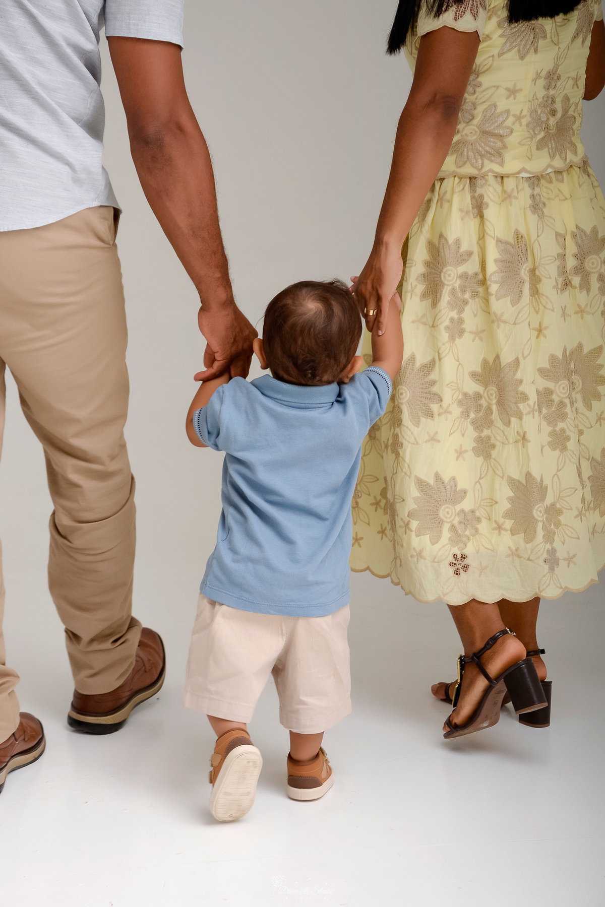Drica studio - ensaio infantil - ensaio de 1 ano - smash the cake - arca de noé - acompanhamneto mensal - mãe e pai segurando as mãozinhas do filho - bebe dando seus primeiros passos - usando camisa azul clara - foto em fundo branco 