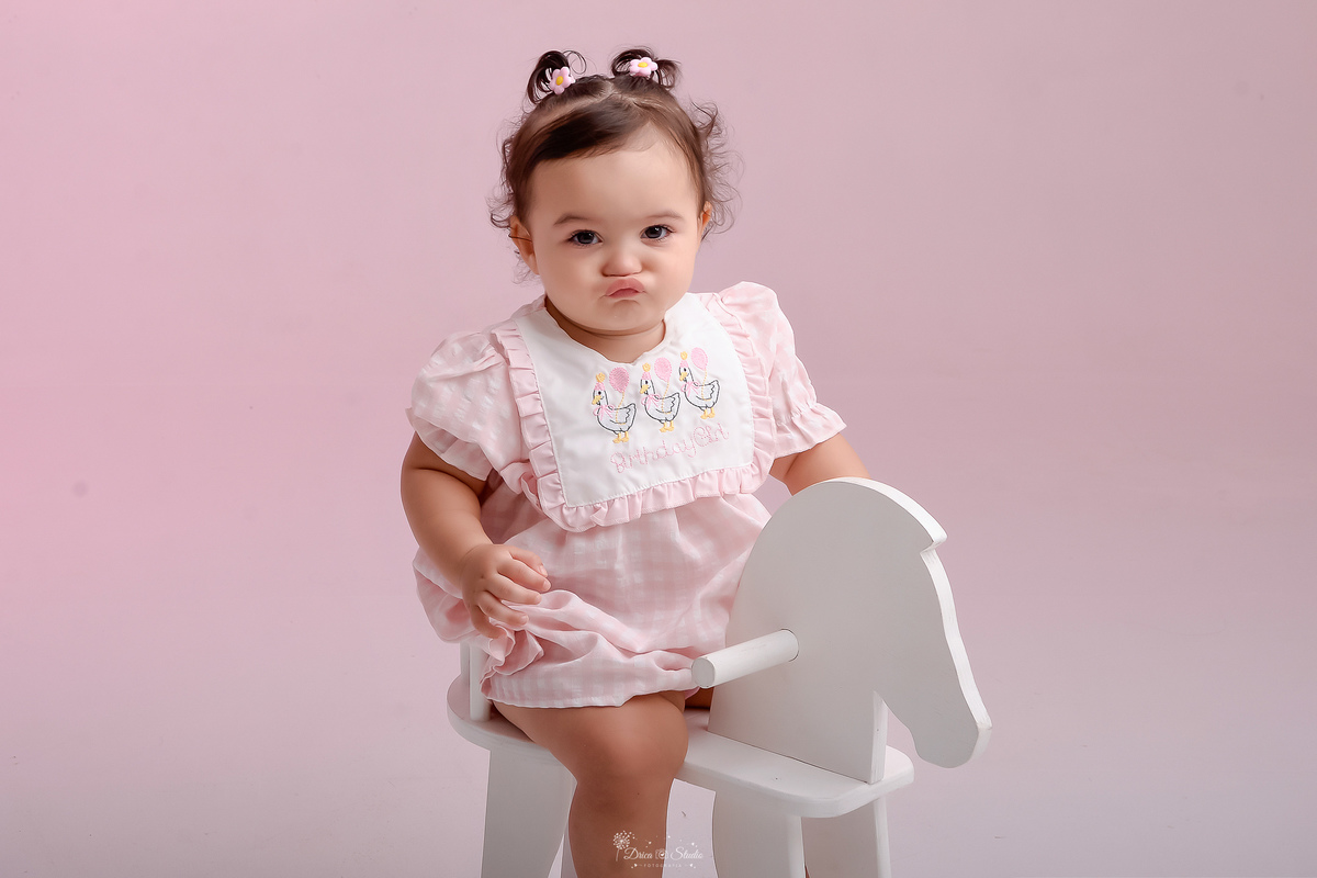Drica studio - smash the cake - ensaio familia - 1 aninho - ensaio em estúdio - bebê sentada em um cavalinho de madeira branco - foto com luz rosa ao fundo - bebe fazendo biquinho 