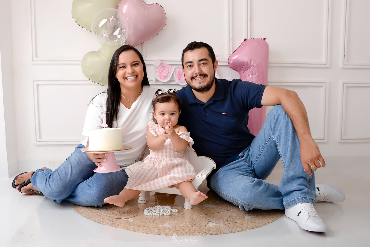 Drica studio - smash the cake - ensaio familia - 1 aninho - ensaio em estúdio - mãe, pai e filha - filha sentada em um baquinho de madeira branco - balões de coração - balão de numero 1 - mãe segurando bolo - bebe batendo palma - one na parede -