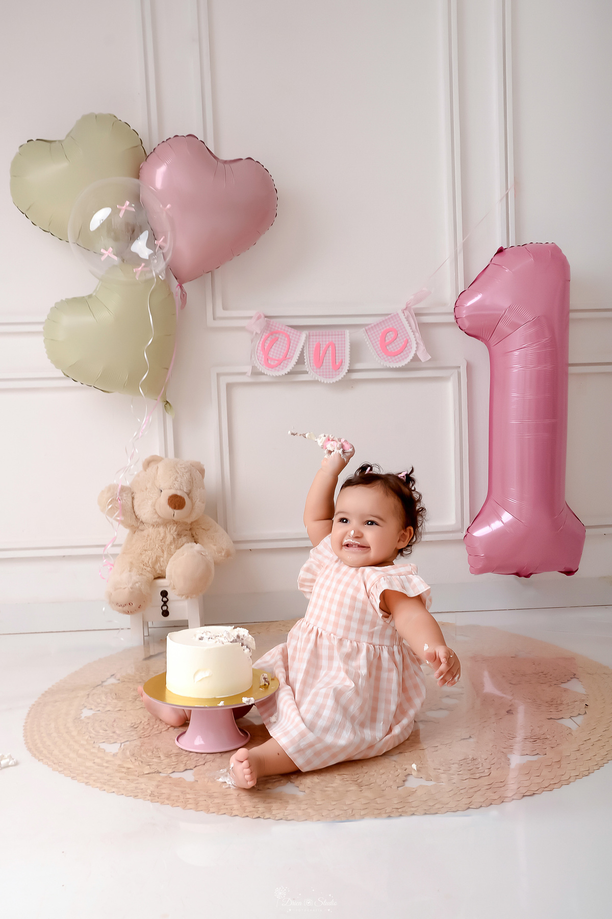 Drica studio - smash the cake - ensaio familia - 1 aninho - ensaio em estúdio - bebe com a mãezinha levantada - ursinho de pelucia ao fundo - balões de coração - balão de numero 1 - bolo branco - parede branca 