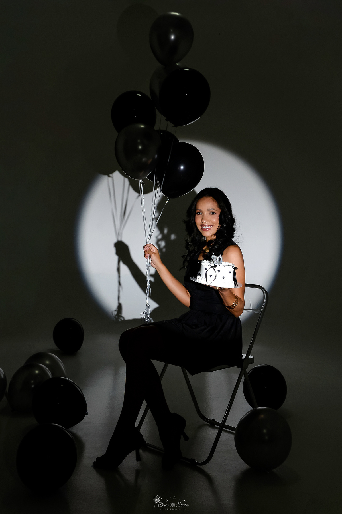 drica studio - xinguara - pará - ensaio de aniversário - 30 anos - trintou - fotos em estudio - sentada em uma cadeira - usando vestido preto - segurando bolo - segurando balões com gas helio na cor preta - balões pretos e pratas pelo chão -