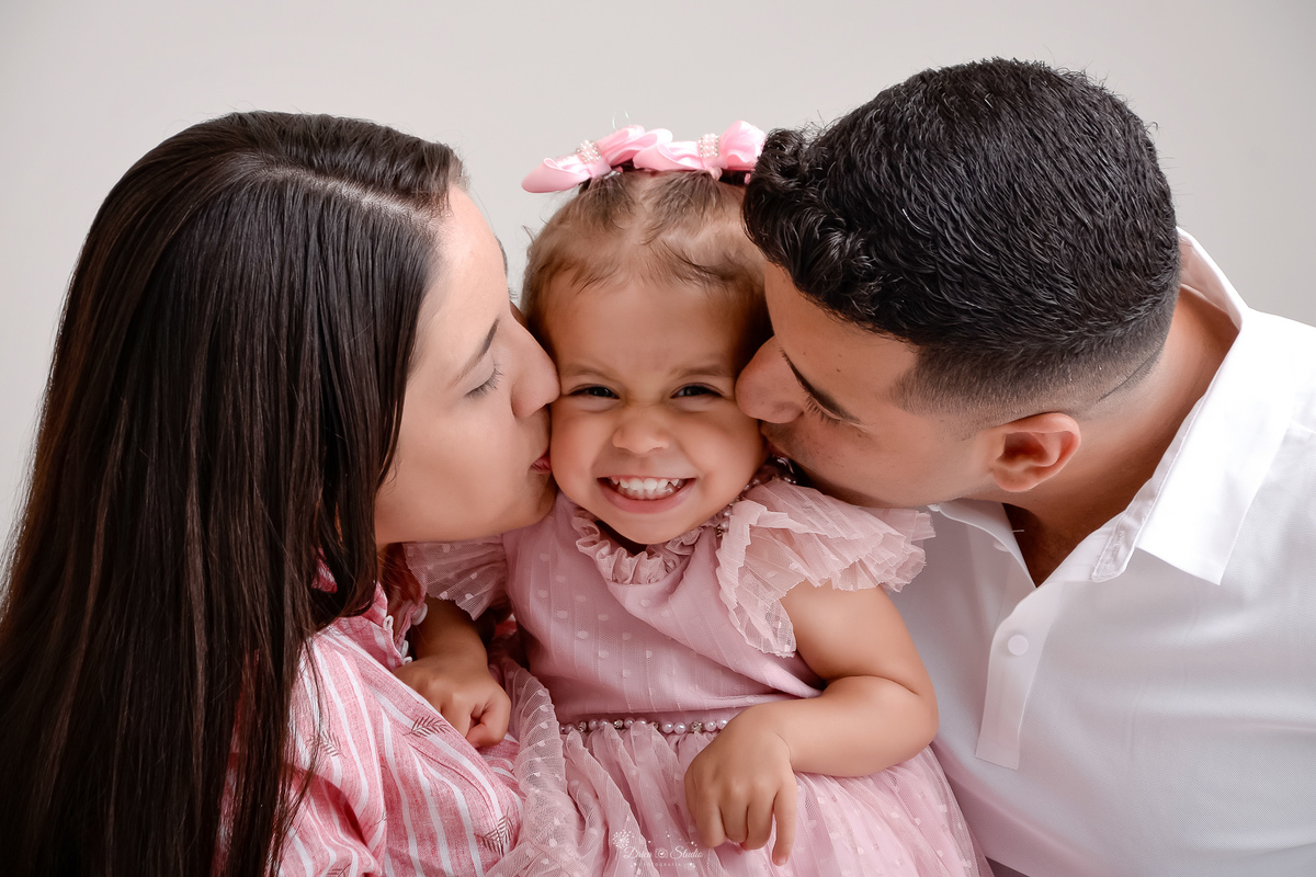 Drica studio - ensaio familia - ensaio de 2 anos - fotos em estudio - mãe, pai e filha - mãe e filha usando vestidos rosas - bebê com laços rosas na cabeça - pai e mãe dando beijinho na filha - foto com fundo branco - pai usando camisa branca 