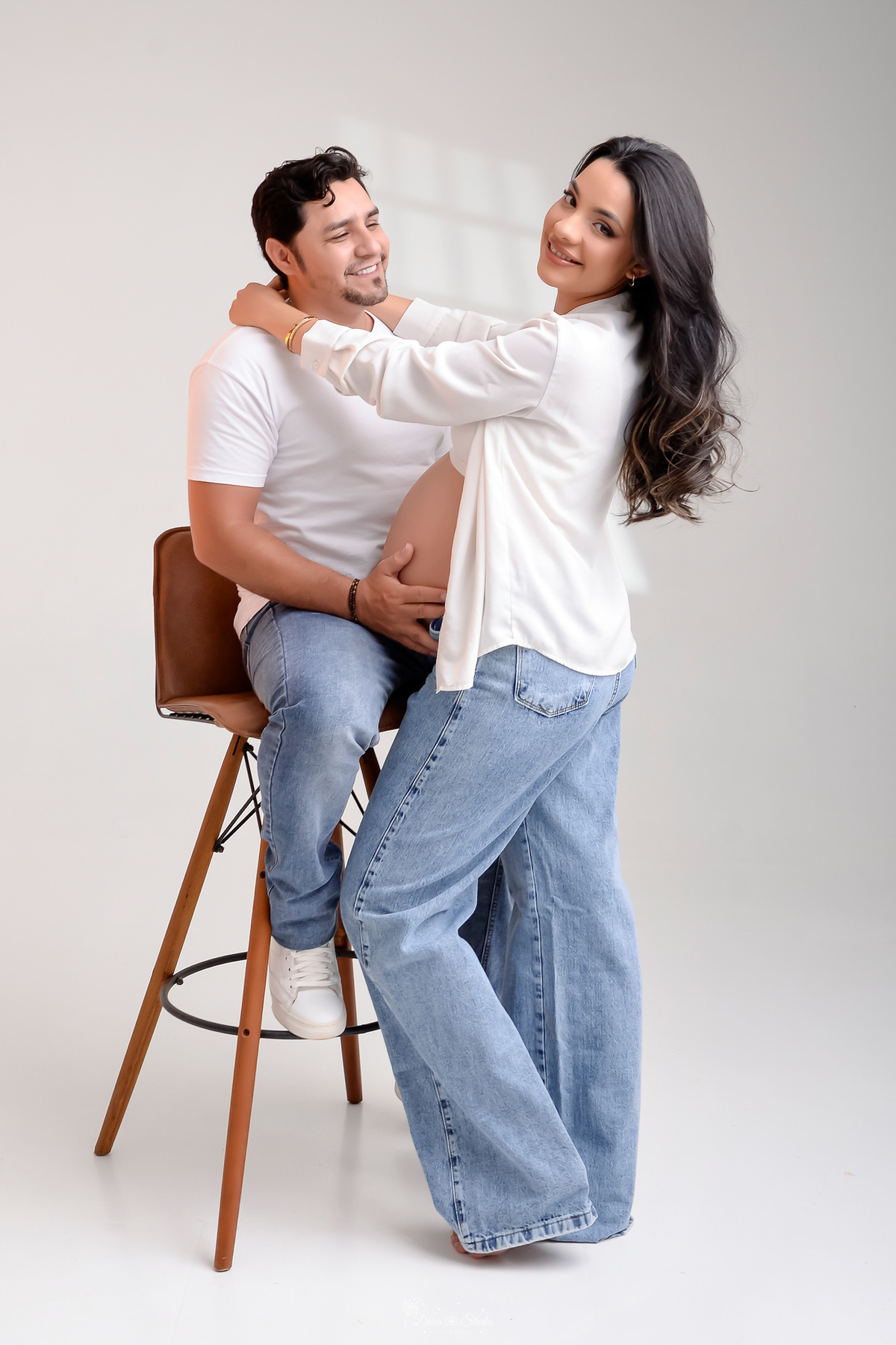 drica studio - xinguara- pará - ensaio gestante - mamãe de primeira viagem - fotografia de gestante - baby boy - papai e mamãe - casal usando blusa branca e calça jeans - pai sentado sobre cadeira - foto com o fundo branco -