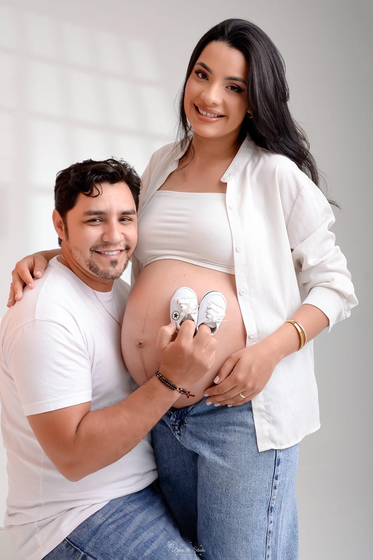 drica studio - xinguara- pará - ensaio gestante - mamãe de primeira viagem - fotografia de gestante - baby boy - papai e mamãe - casal usando blusas brancas e calça jeans - pai segurando sapatinho sobre a barriga da mamãe - foto em fundo branco - 