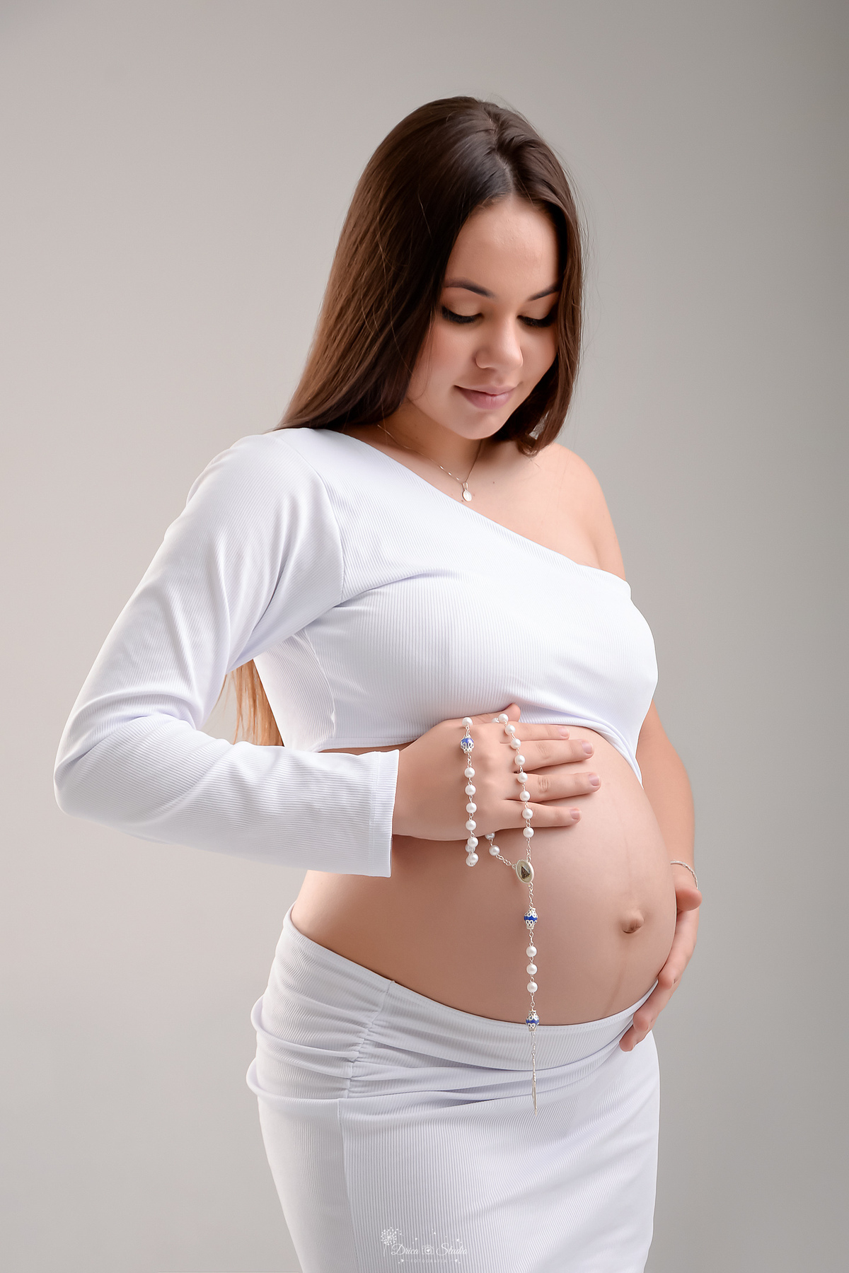 Ensaio gestante - ensaio em estudio - mamãe de primeira viagem - drica studio - xinguara - pará - mamãe usando vestido branco - com as mãos e um terço sobre a barriga - foto em fundo branco 