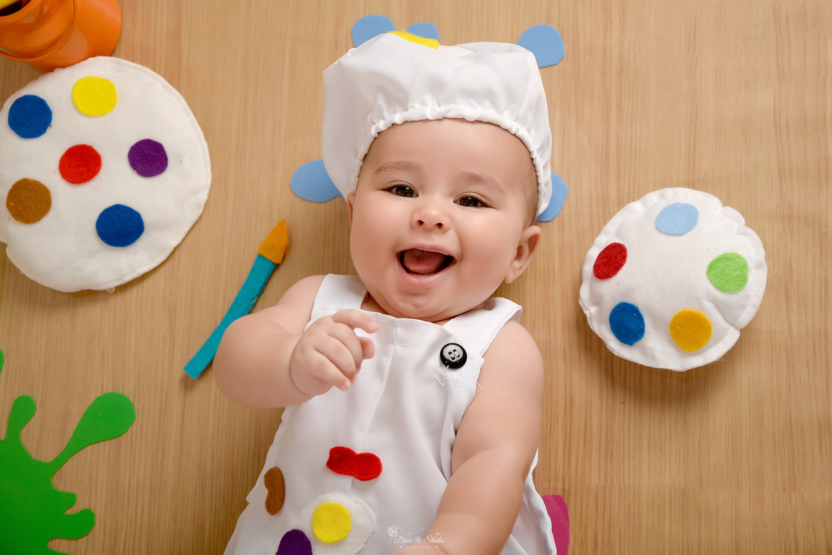 acompanhamento infantil - cenário de aquarela - dia das crianças - mês das crianças - cenários com latas de tintas - bebe deitado no cenário - ao lado de tintas e pincéis de pintura - usando roupinha de pintura e gorrinho na cabeça - 