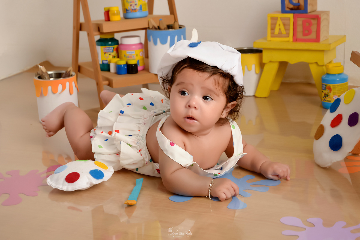acompanhamento infantil - cenário de aquarela - dia das crianças - mês das crianças - cenários com latas de tintas - bebe deitada no cenário - marcas coloridas de tintas pelo chão - bebe usando roupinha de pintura com gorrinho na cabeça - 