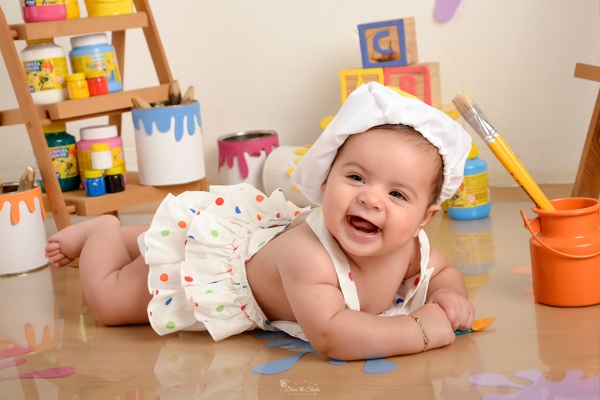 acompanhamento infantil - cenário de aquarela - dia das crianças - mês das crianças - cenários com latas de tintas - bebe usando roupinha de pintora com gorrinho na cabeça - deitada no cenário e sorrindo para a foto - 