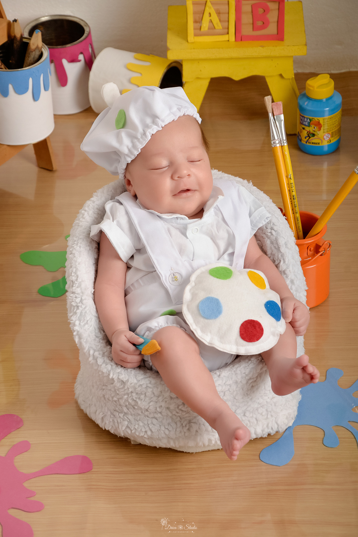 acompanhamento infantil - cenário de aquarela - dia das crianças - mês das crianças - cenários com latas de tintas - bebe dormindo sobre sofazinho - usando roupinha de pintor e gorrinho na cabeça - segurando um pincel de pintura 