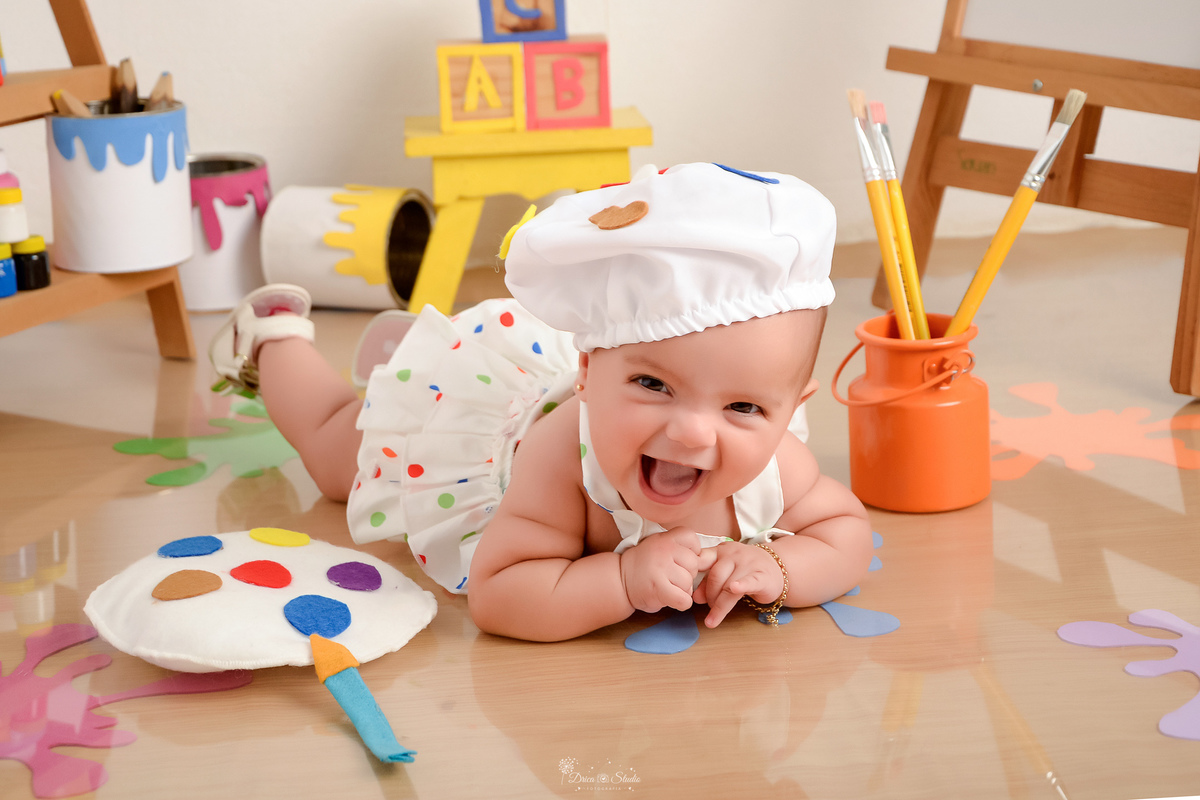 acompanhamento infantil - cenário de aquarela - dia das crianças - mês das crianças - cenários com latas de tintas - bebe deitada no cenário - ao lado de lata de tinta com pincéis - usando roupinha de pintora - com gorrinho na cabeça - sorrindo para a fot