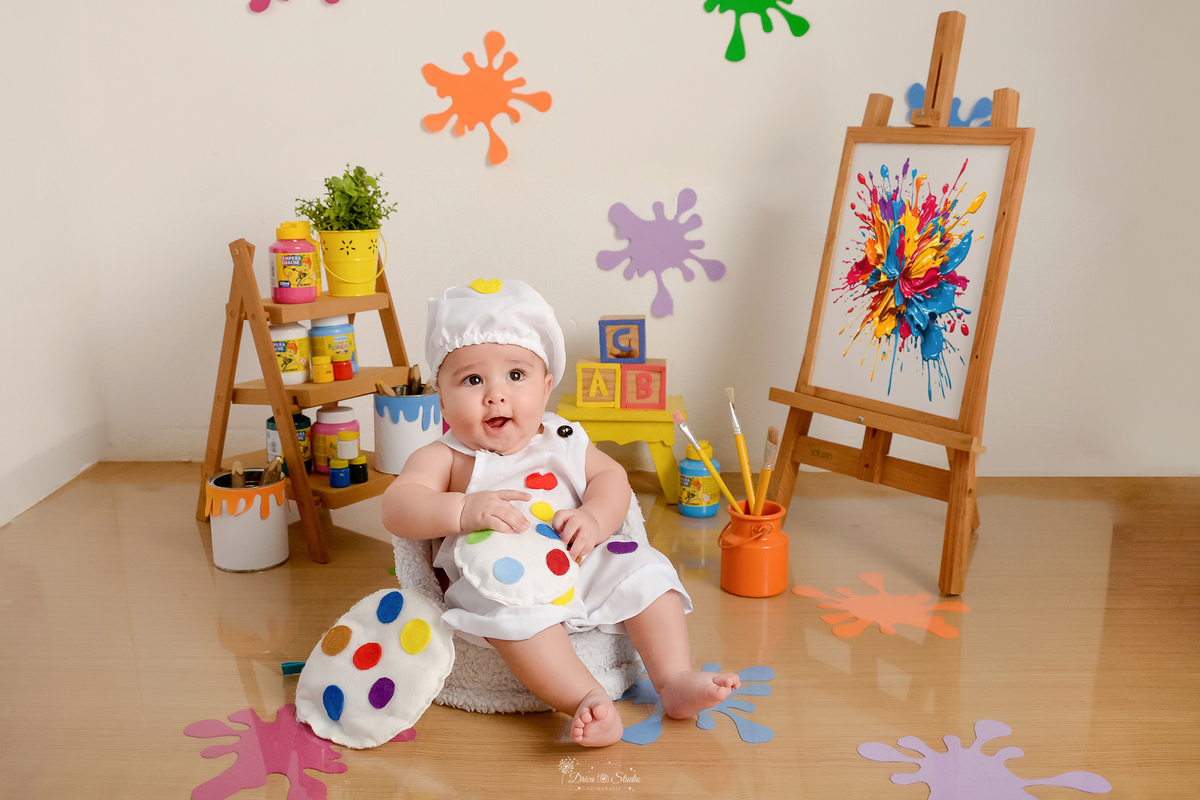 acompanhamento infantil - cenário de aquarela - dia das crianças - mês das crianças - cenários com latas de tintas - bebe sentado em mini sofazinho - marcas de tinta na parede e no chão - lousa com pintura - bebe usando roupinha de pintor e gorrinho 